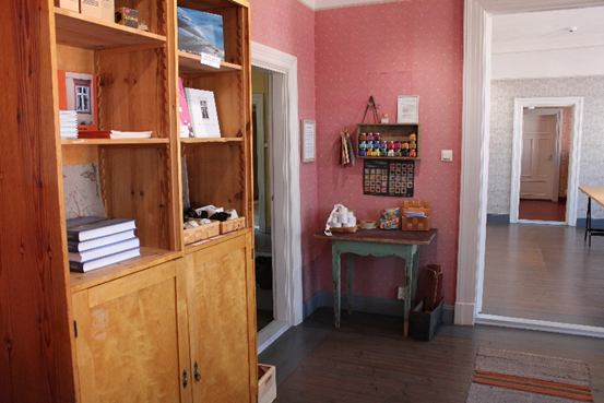 kuva huoneen nurkasta, takana oikealla avautuu oviaukko toiseen huoneeseen, etualalla vasemmalla puinen kirjahylly. Kirjahyllyn vieressä oviaukko vasemmalle. Kuvan keskellä, huoneen nurkassa on vanha pöytä ja seinässä hyllykkö.