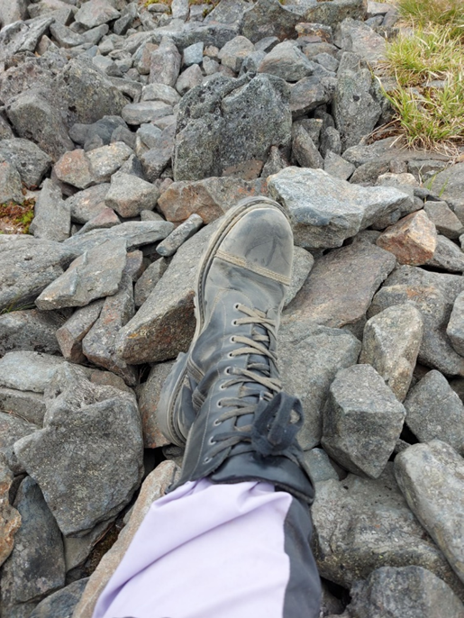 Kuvassa keskellä jalka, jalassa musta, hiekkapölystä harmaantunut maihinnousukenkä. Jalan alla kiviä.