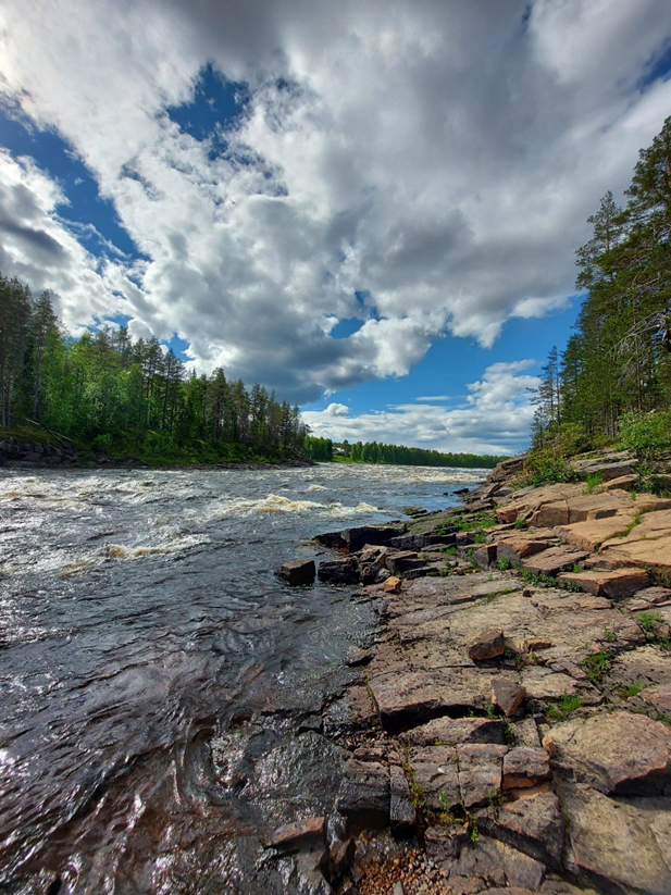 Kuvassa keskeltä vasempaan alareunaan leviävä joki. Kuvan yläreunassa sininen, pilvinen taivas. Kuvan oikealla reunalla kallioita ja metsää.