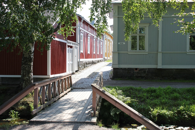 Kuvassa keskellä pieni puusilta, joka johtaa mukulakivillä päällystetylle kadulle. Katu kulkee kuvan keskellä. Kadun vasemmalla puolella on punainen puutalo ja oikealla reunalla on vaaleansininen puutalo.