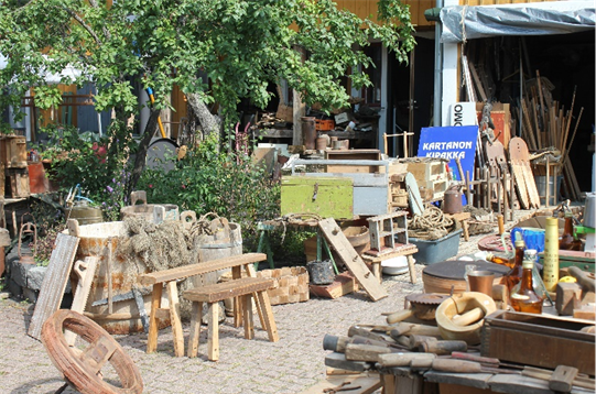 Kuva erään pihakirppiksen tarjonnasta. Kuvassa hieman vasemmalla omenapuu, jonka ympäsrillä paljon vanhoja, myynnissä olevia esineitä kuten pärekoreja, kiuluja ja puisia jakkaroita. Tavaroiden vieressä mukulakivillä päällystetty kulkuväylä, joka johtaa autotalliin. Kulkuväylän oikealla puolella on lisää myytäviä tavaroita.