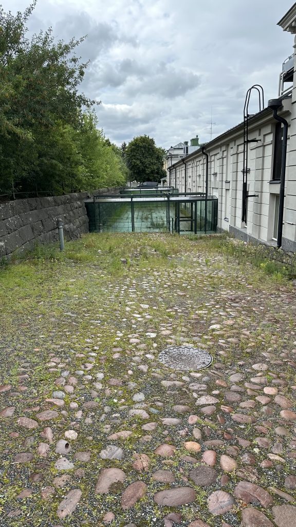 Backyard of the museum. Museum building on the right and old stone wall on the left. Greenish paving and the glass corridors in the middle.