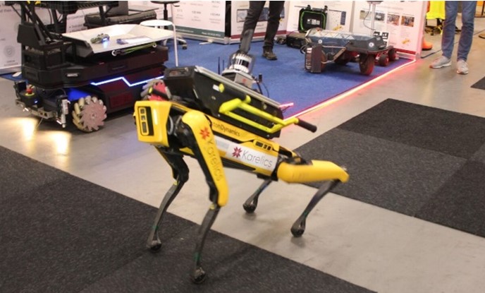 Etualalla Spot-robottikoira ja taustalla erilaisia robotteja sekä niiden toimintaa ja ohjausta esittelevä messuosasto.