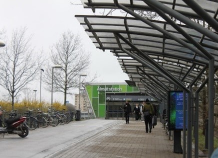 Älvsjön juna-aseman sisäänkäynti ulkoa päin kuvattuna. Oikealla asemalle johtava katettu kävelytie ja vasemmalla pyöriä pyörätelineessä.
