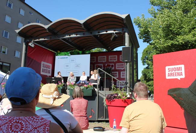 Näkymä katsomosta Eetunaukion lavalle, jossa neljä panelistia ja juontaja käyvät keskustelua.