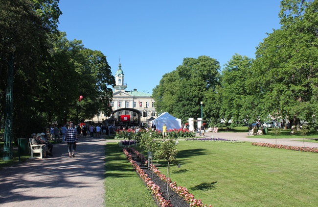 Näkymä Raatihuoneenpuistosta. Etualalla nurmialue ja hiekkapolut, reunoilla suuria lehtipuita ja taustalla esiintymislava sekä Raatihuone. 